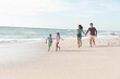© wavebreak3 - Full length of multiracial parents following behind children running on shore at beach against sky