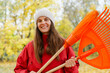 © sementsova321 - A woman stands in a park holding orange rakes while smiling. Yellow leaves fill the trees around her on a clear autumn day