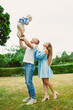 © DSMT - Family enjoys a sunny day in the park, with a father lifting his child into the air while the mother smiles and watches from below, all in a green outdoor space