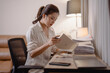 © MINAE - Young Asian woman concentrating over a desk stacked with paperwork and files, reviewing documents and organizing a busy home office workload for work or study