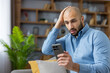 © Liubomir - Upset young man in a blue shirt sitting on a sofa at home, looking shocked and frustrated while reading disturbing information on his smartphone with a hand on his head