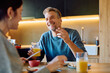 © we.bond.creations - Happy man and woman having breakfast together, laughing while drinking orange juice and talking in a modern kitchen