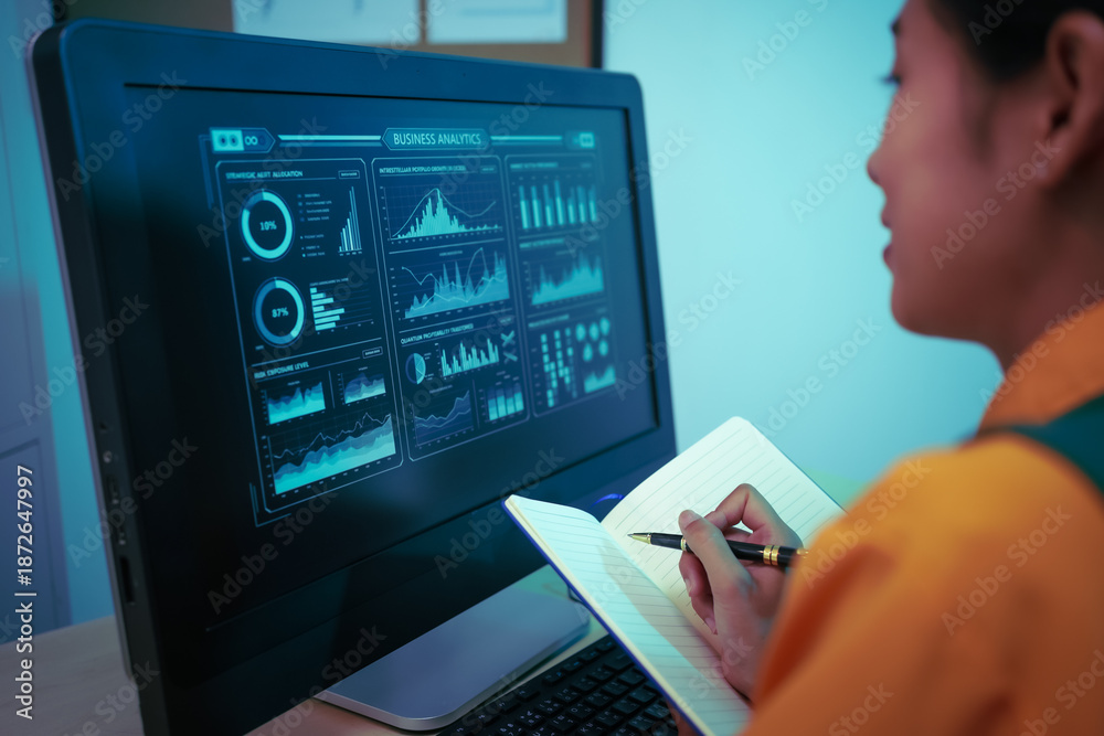 Analyst reviewing digital dashboard of charts and graphs on large computer monitor, taking notes on business intelligence, data analytics, KPI performance metrics and financial trends

