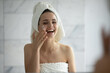 © fizkes - Morning hydration fun. Head shot laughing joyful young lady in towel turban on head applying tender moisturizing cream dots on facial skin after bath enjoying home spa beauty skincare ritual by mirror