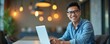 © Vadym - Smiling asian man works on laptop in bright office. Wears eyeglasses, blue shirt. Freelancer student uses computer for work. Modern workplace interior, tech lighting concept. Cheerful employee poses