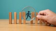 © Norah Allen - A magnifying glass examines a running silhouette figure positioned between wooden blocks on a light wood surface against a teal backdrop business concept person search looking career
