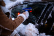 © Alliance - A person in warm clothing uses a blue tool to scrape ice and snow off a car windshield at night, with city lights blurred in the background.