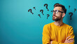 © miss irine - Man with glasses wears yellow sweater arms crossed, looking pensive with drawn question marks around his head. He appears confused, pondering life choices or seeking answers. Blue background.