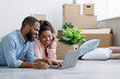© Prostock-studio - Smiling millennial black couple rest, lie on floor with cardboard boxes with things and plants in room and look at laptop. Furniture purchase, planning interior and online shopping, design create