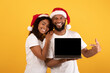 © Prostock-studio - Christmas sale. Excited african american spouses showing laptop with blank screen, pointing at it, posing in Santa hats and smiling at camera over yellow background