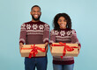 © Prostock-studio - Happy african american couple stretching Christmas gifts to camera over blue studio background. Excited spouses wearing winter sweaters and celebrating New Year 2022 together