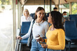 © Prostock-studio - Public Transportaton. Two smiling young women standing at autobus, talking and looking at each other, holding takeaway coffee cup and cell phone, students enjoying ride or travel together in vehicle