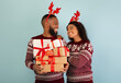 © Prostock-studio - New Year celebration. Loving african american spouses in sweaters and deer horns, man holding heap of Christmas gifts over blue background, studio shot