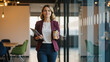 © ViskBx - Confident businesswoman walking through an office corridor with coffee and tablet in hand