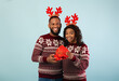 © Prostock-studio - Loving african american man giving gift box to happy wife and celebrating New Year or Christmas together over blue studio background. Happy black couple wearing deer horns