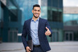 © Prostock-studio - Positive millennial arab man in formal stylish suit outstretching hand towards camera and smiling, greeting business partner next to office building, holding modern laptop, copy space