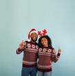© Prostock-studio - Look here. Happy african american couple in Christmas outfits pointing up at free space, woman wearing deer horns and man posing in Santa hat over blue background, crop
