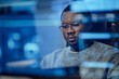 © Seventyfour - Black man young adult wearing glasses sitting at desk working on computer with digital interface reflections visible on screen, focusing on technology task in modern workspace