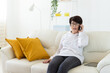 © satura_ - Woman talking on smartphone on white sofa in modern living room. Home communication, remote conversation, and digital lifestyle inside bright apartment interior.
