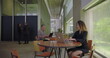 © Marco - Young woman working on laptop at wooden table in bright modern office lounge with large windows and colleagues in background, everyday business environment