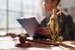 © Tj - Lady Justice statue and a wooden gavel on a table, with a lawyer reviewing documents in the background, symbolizing legal proceedings and judicial system