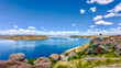 © Travel 'n' Lifestyle - View of bright blue waters under a vast sky with fluffy clouds, contrasted by the rugged, rocky terrain and greenery, Sillustani, Peru.