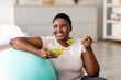 © Prostock-studio - Sports and healthy nutrition for weight loss concept. Curvy black lady leaning on fitness ball, eating fresh vegetable salad at home. Plus size young woman enjoying slimming and exercising program