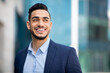 © Prostock-studio - Closeup portrait of handsome young bearded arab man in suit entrepreneur or manager posing by modern business center, smiling and looking at copy space. Business opportunities concept