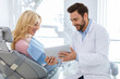 © Prostock-studio - Smiling man dentist showing female patient jaw x-ray on digital tablet, dental clinic interior. Bearded man stomatologist having conversation with lady in dental chair, holding pad, side view