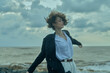 © SHOTPRIME STUDIO - Wind, beach, ocean, movement, coat, shirt frame a windy scene as a woman on the shore stretches arms, faces the sea, and basks in the dramatic sky with rolling waves and misty light