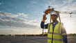 © Vyatcheslav - Smiling male construction worker in safety vest and hard hat making OK sign at sunset. Black engineer at industrial site with crane. Workplace safety and success concept. Copy space