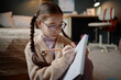 © Seventyfour - Caucasian girl child with long brown hair in braids wearing glasses sitting on floor holding pencil and writing in notebook, showing focused expression in home setting