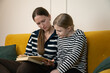 © yaroslav1986 - Mother and daughter sitting together on a yellow sofa in their living room, bonding while reading an open book, emphasizing family time, education, and childhood learning indoors