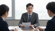 © Sterling - A man in a gray suit sits at a conference table holding documents, speaking to two colleagues across from him in a bright office with large windows