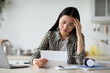 © Prostock-studio - Upset young asian woman sitting at table with modern laptop in kitchen, reading letter and touching her forehead, copy space. Sad lady holding document or bills, got bad news, home interior