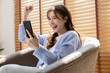 © 220 Selfmade studio - Excited young asian woman cheering while holding smartphone sitting on cozy sofa indoors showing winning expression and success feeling