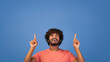© Prostock-studio - A young man with curly hair smiles widely while pointing both of his index fingers up. He wears a light orange shirt. The background is a solid blue color.