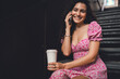© BullRun - Hispanic woman in floral dress smiles warmly during smartphone call while sitting with coffee, symbolizing mobile lifestyle, approachable professionalism, and joyful digital connection.