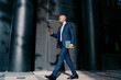 © BullRun - Focused businessman in tailored suit walking confidently in urban business district, multitasking with smartphone and digital tablet, symbolizing mobile decision-making and digital leadership.