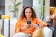 © Anastasiya - Smiling adult caucasian woman sitting in hotel lobby using smartphone near yellow suitcase, relaxed travel lifestyle moment with modern interior, comfort and mobile communication