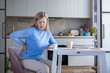 © Liubomir - Senior woman sitting at her kitchen table, wincing and holding her lower back in pain, illustrating chronic back or kidney discomfort, aging, fatigue and home care struggles