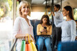 © Prostock-studio - Public Transportation. Group of diverse people taking bus, selective focus on smiling mature woman holding shopping bags, standing and looking out of window, leaning on handle going home