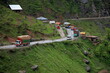 © Travel 'n' Lifestyle - View of trucks and vehicles navigating a winding mountain road amidst lush green slopes and scattered buildings, Lowari Pass, Khyber Pakhtunkhwa, Pakistan.