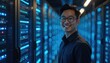 © miss irine - Asian man smiles in data center with rows of servers glowing blue. IT specialist works with computer hardware. Tech infrastructure is operational and illuminated.