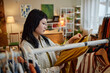 © Seventyfour - Asian young adult woman browsing second hand clothing rack, examining garment closely in thrift store setting, surrounded by various clothes, focused on sustainable shopping