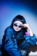 © Seventyfour - Portrait of young adult Asian woman sitting on floor adjusting sunglasses with confident expression, wearing second hand clothing, looking directly at camera in studio setting