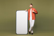 © ViDi Studio - Full size young smiling happy man wear casual clothes stand at big huge blank screen mobile cell phone smartphone with workspace copy space mockup area isolated on green background. Lifestyle concept