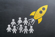 © Daria - Conceptual illustration of a yellow rocket launching from a group of white chalk-drawn people symbolizing innovation and leadership on a blackboard background