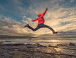 © mark_gusev - A man in a red jacket jumps high on a rough stone beach, with the sun setting in the background. Scene is peaceful and serene, as the man is enjoying the beauty of the natural surroundings