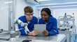 © SYATRI RAWU - Two young engineers in safety goggles and blue overalls examining a tablet near a robotic arm in a bright laboratory setting, discussing technology